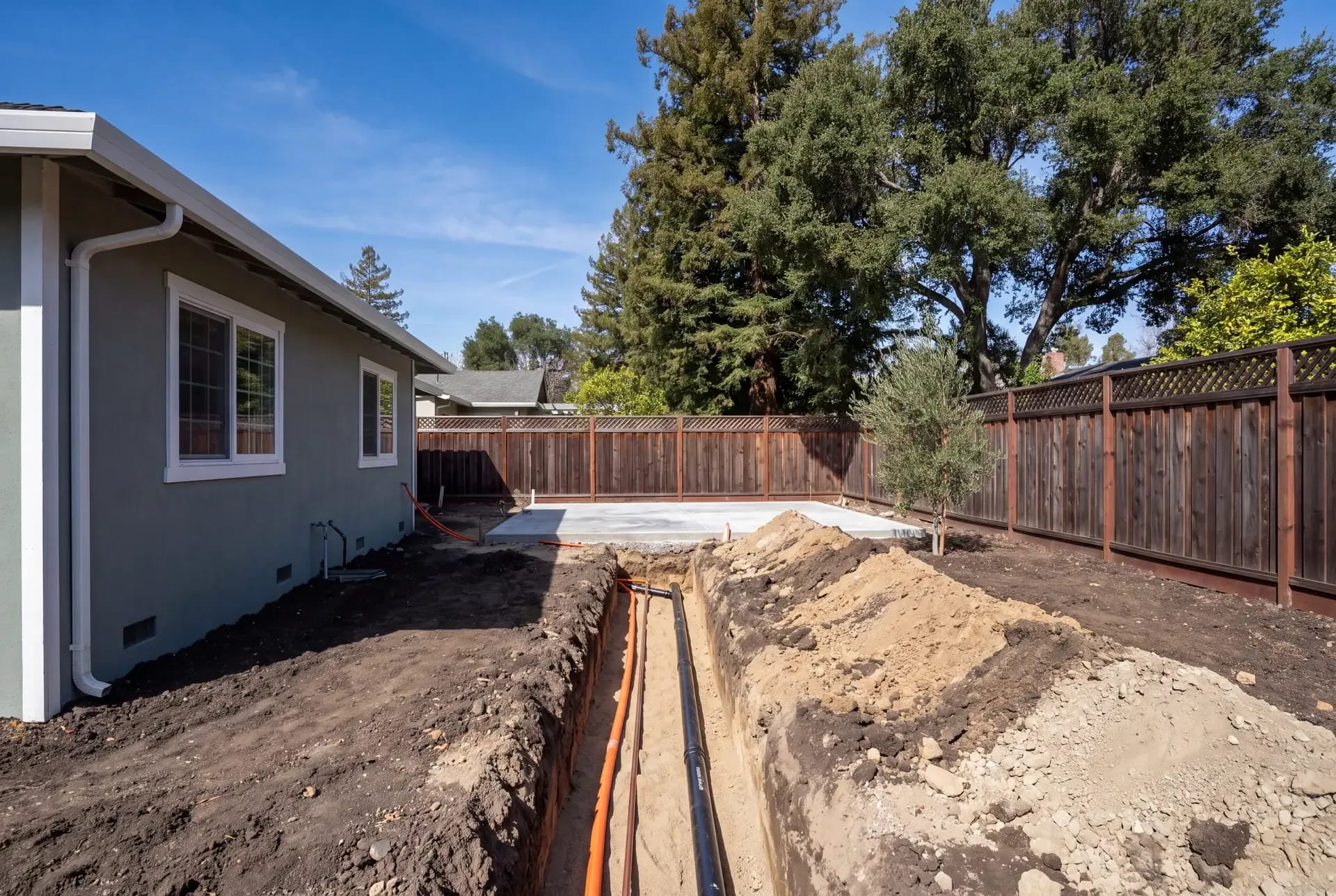 An open utility trench from a main house toward a freshly poured ADU slab, with electrical, water, and sewer lines at depth