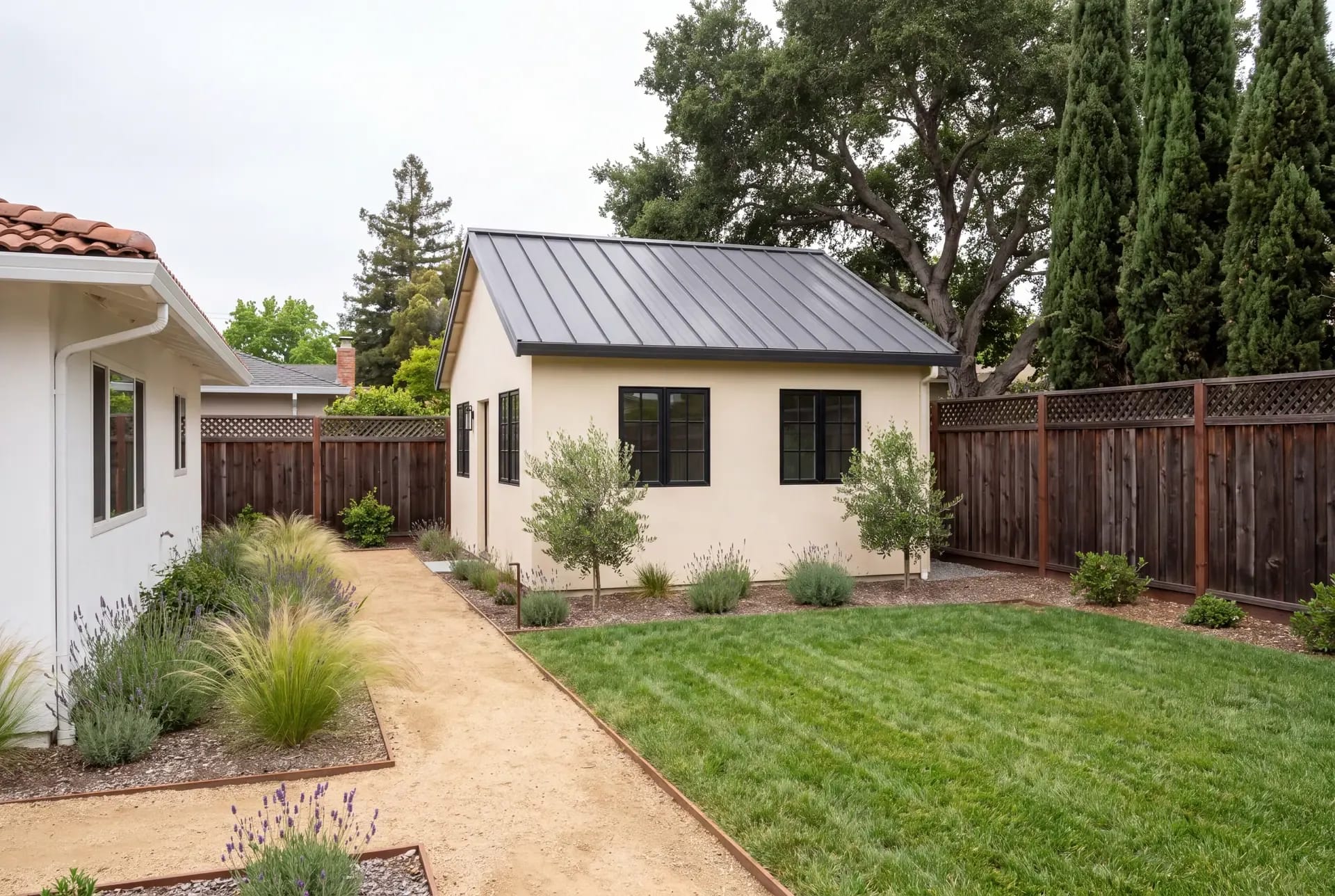 A newly completed detached ADU in a Bay Area backyard with a planted path from the main house, redwood fence and mature coast live oak behind