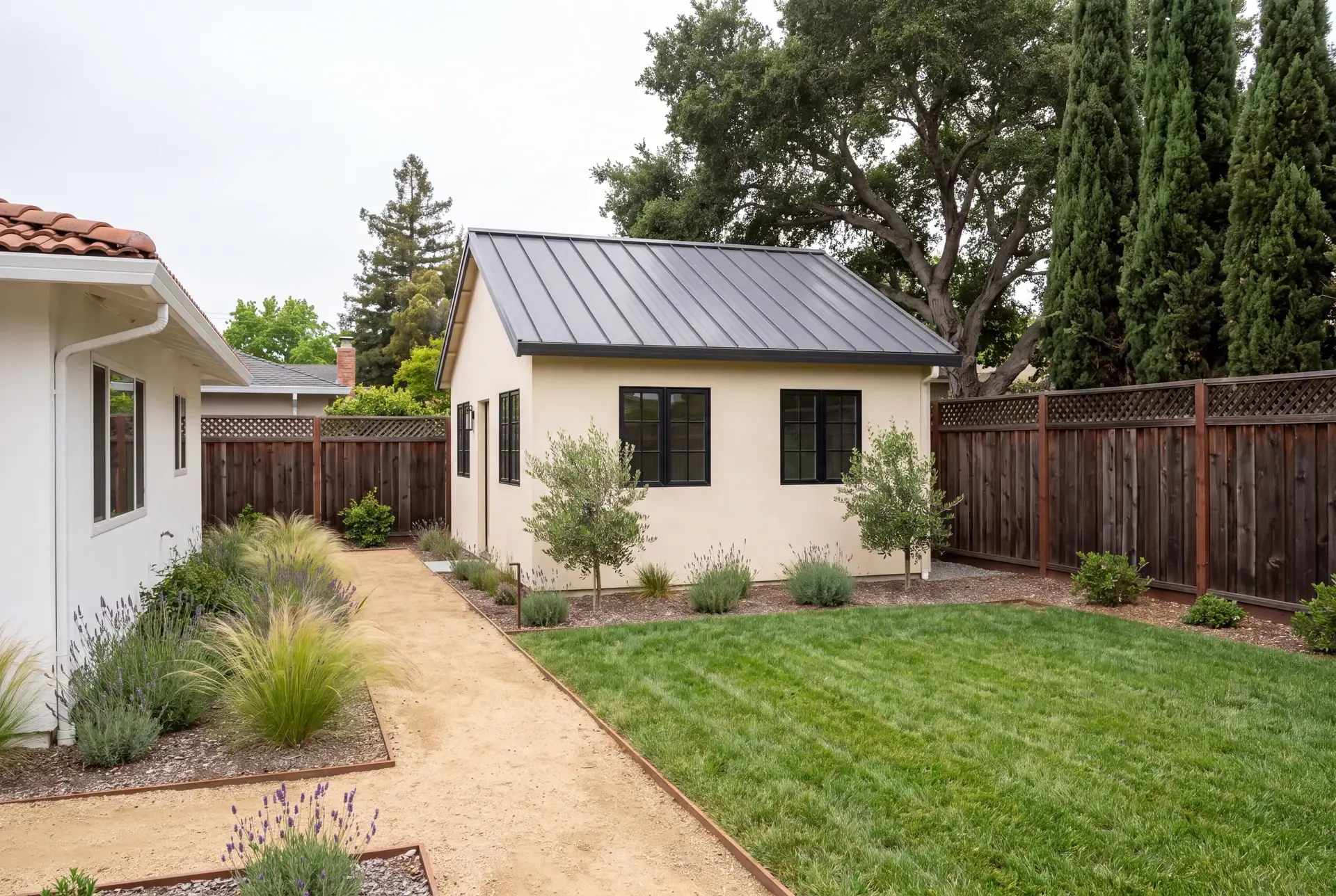 A newly completed detached ADU in a Bay Area backyard with a planted path from the main house, redwood fence and mature coast live oak behind