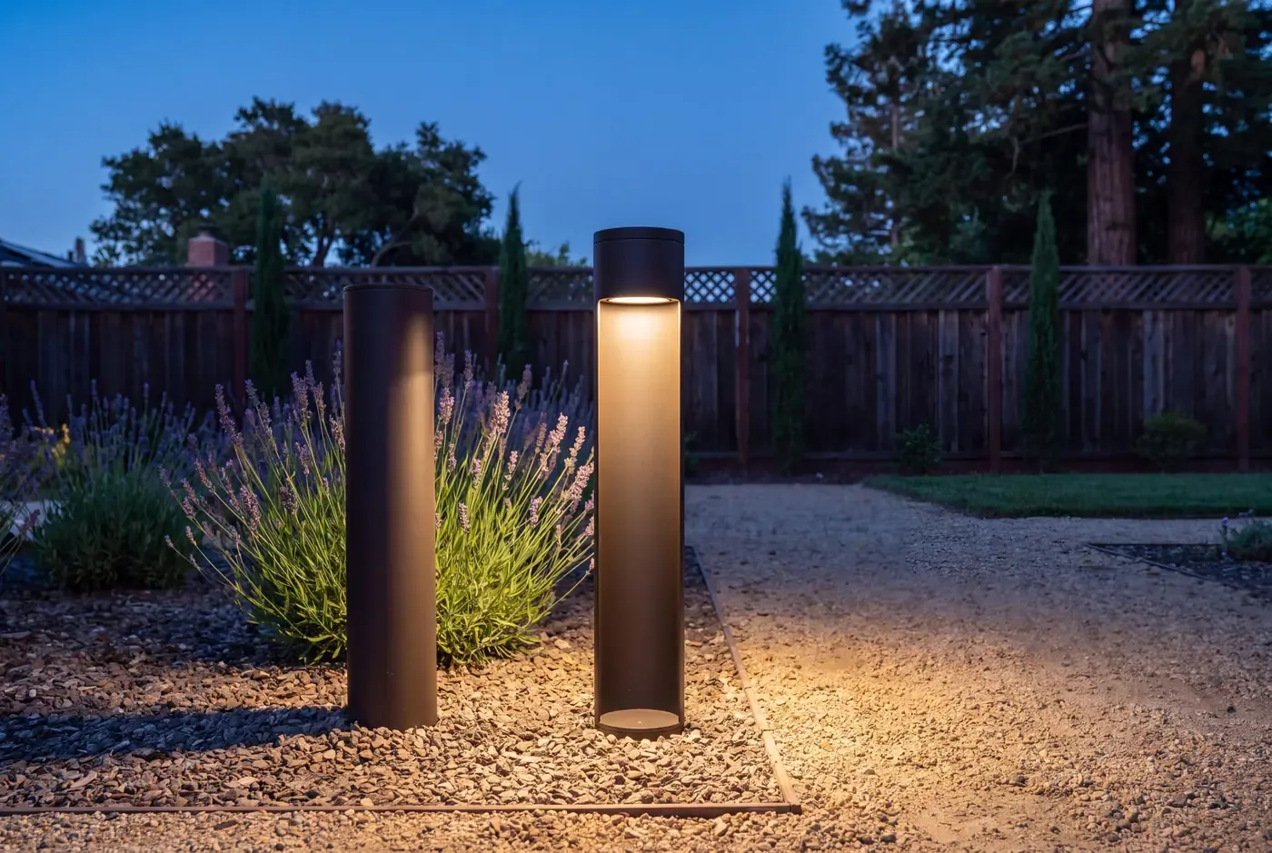 A dark bronze cylindrical path light in a gravel bed at dusk, downward cone of warm light on adjacent plants