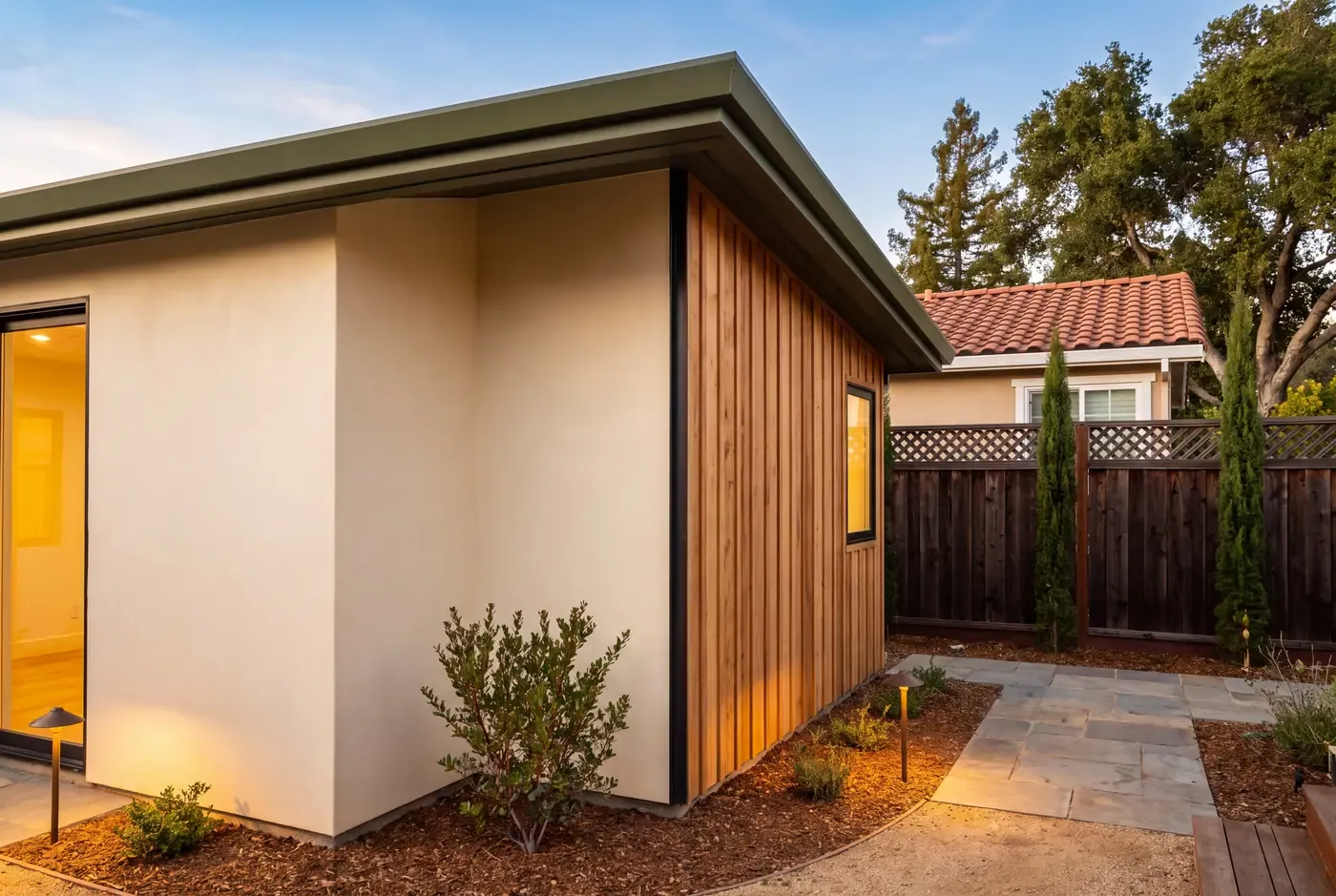 Exterior corner of a newly built ADU where smooth stucco transitions to vertical cedar siding at a blackened steel flashing detail