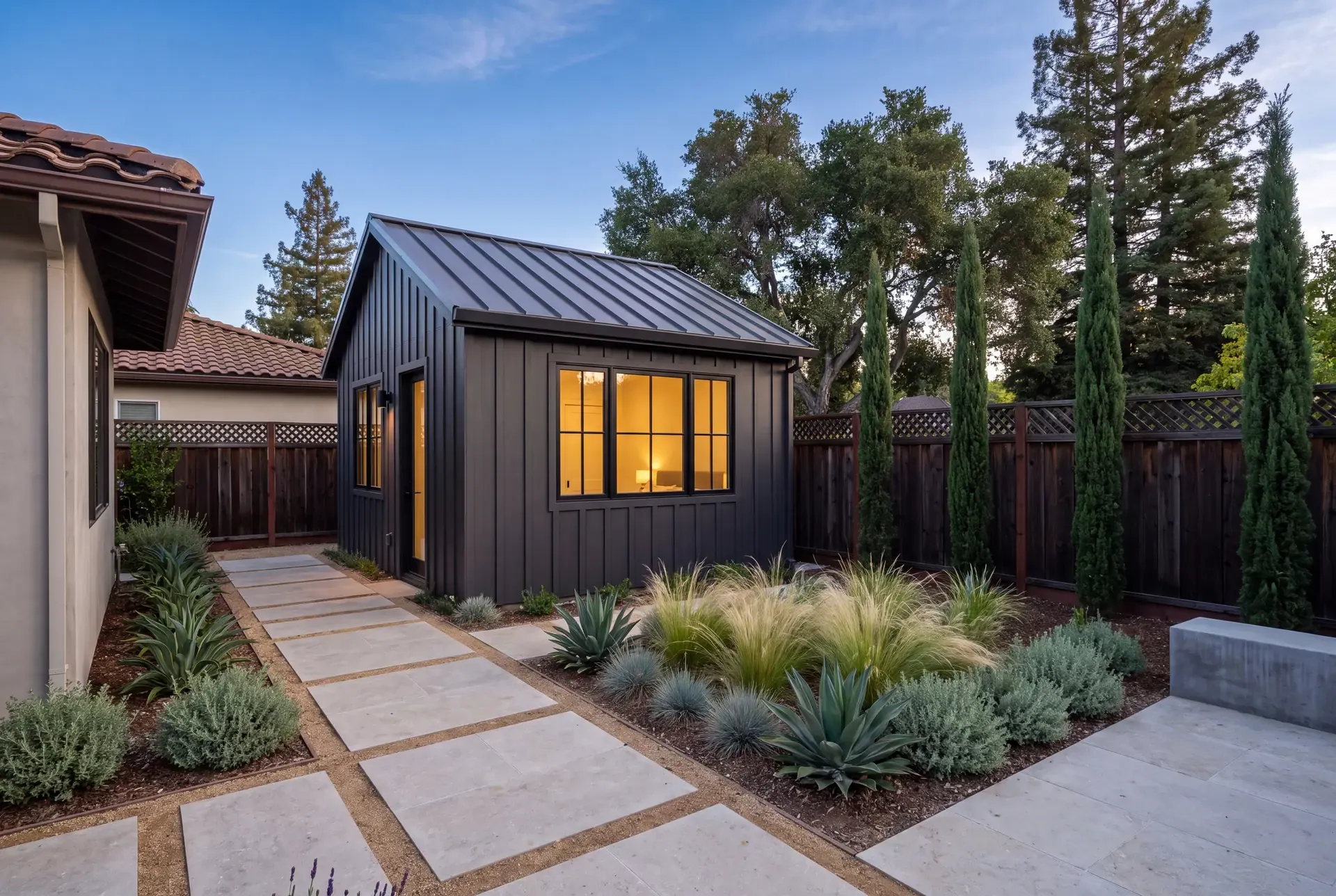A newly completed detached ADU at dusk with vertical board-and-batten siding, black-framed windows glowing warm, a limestone paver path through agave and feather grass