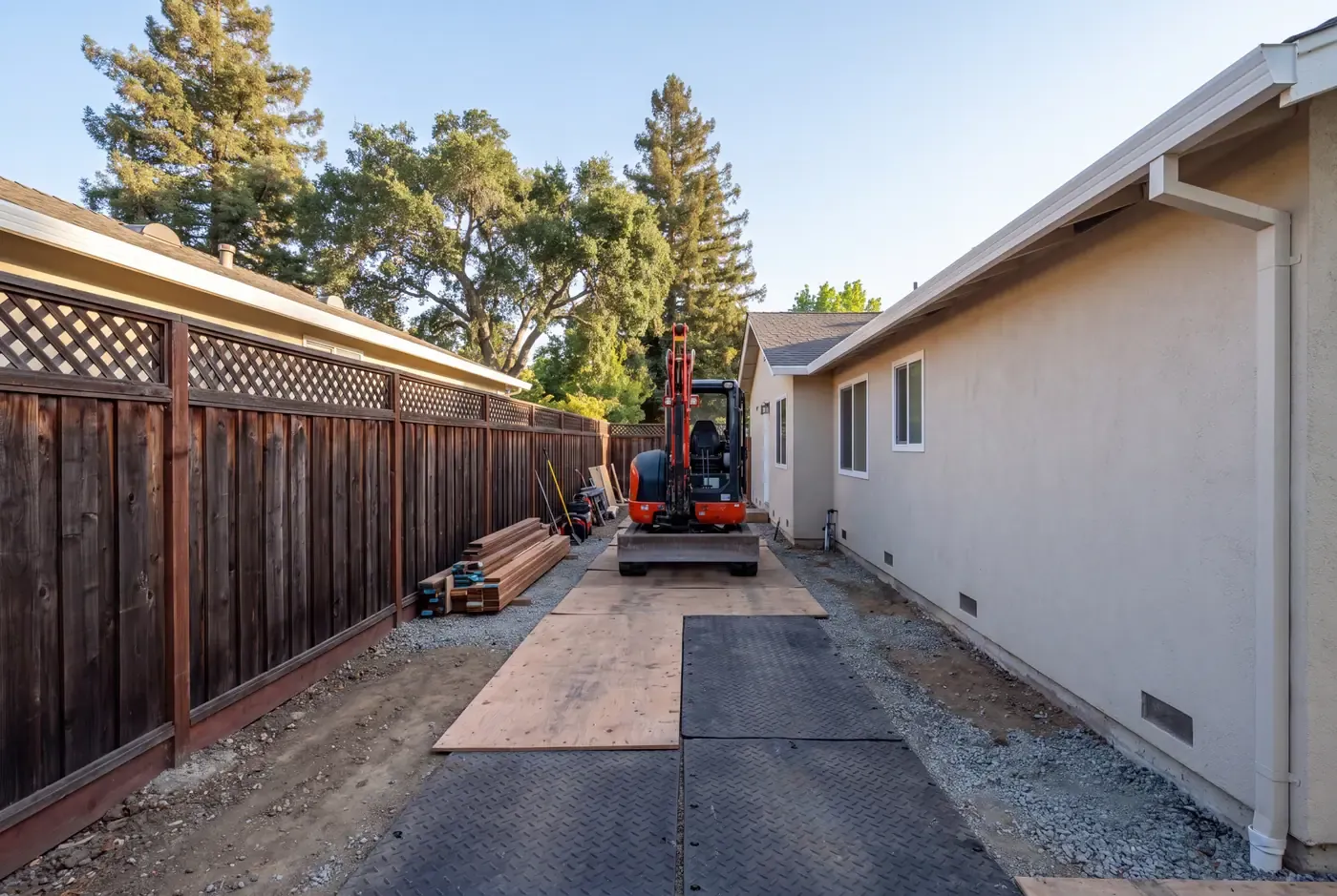 A compact mini-excavator passes through a narrow Peninsula side yard on plywood ground protection